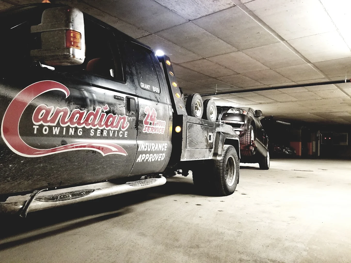 Tow truck with a car on a flatbed inside a dim parking garage, Canadian Towing Service branding visible on the side