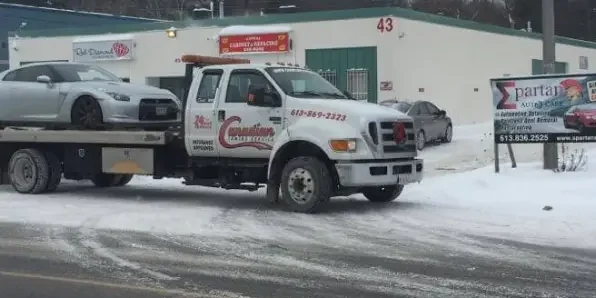 Tow truck hauling a silver sports car on a snowy street, parked in front of a white building with green trim and the number 43.