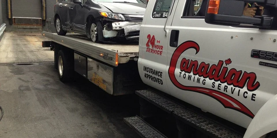 Tow truck with a damaged gray sedan loaded on a flatbed, parked at a service bay. The truck features a red Canadian Towing Service logo and 24-hour service text on the door.