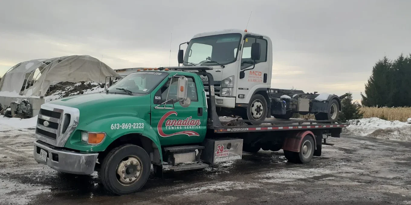 Green Canadian Towing Service tow truck carrying a white semi on a flatbed in a snowy, muddy lot.
