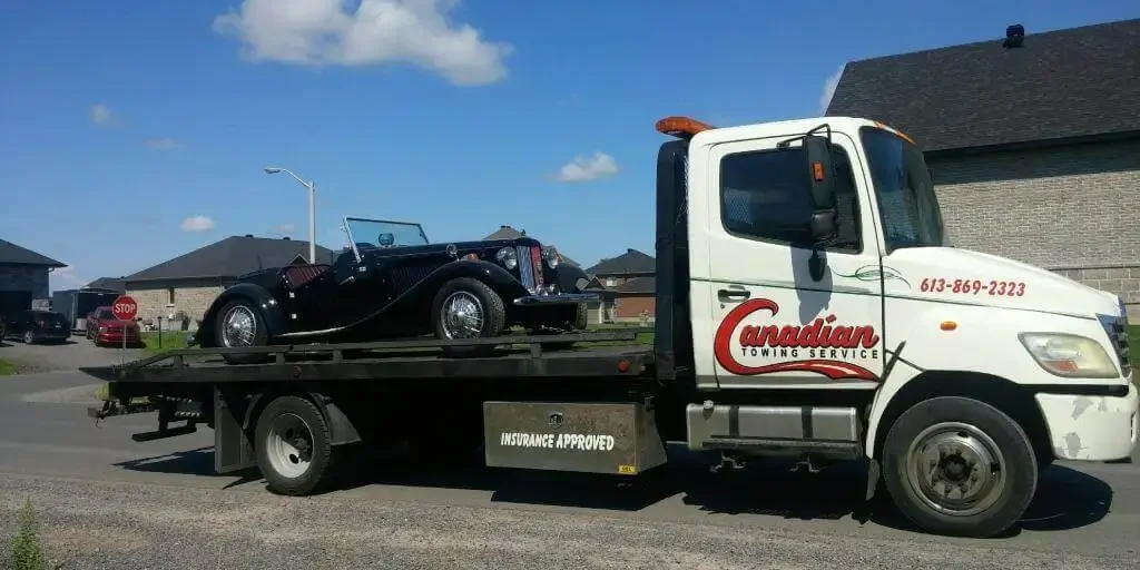 Tow truck labeled Canadian Towing Service carries a classic black convertible on a flatbed in a suburban street attack.